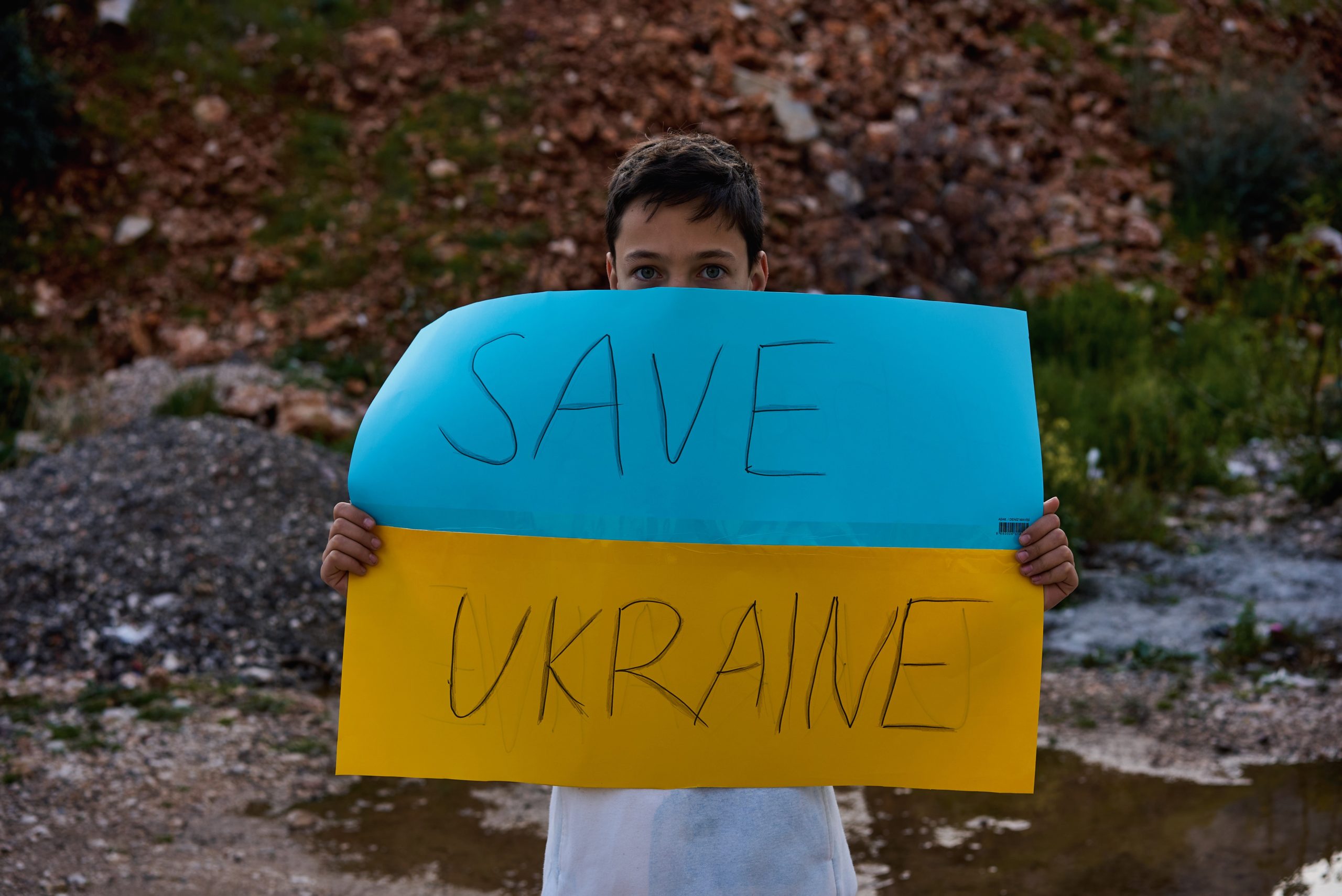 Portrait of Ukrainian boy child near the destroyed building showing banner with massage text Save Ukraine. Asking for help. Crisis, war, no peace, stop aggression from Russia country